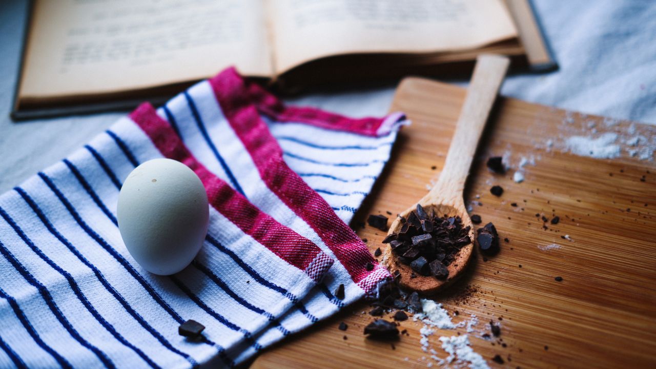 gallery-04 Overhead view of baking essentials with egg, chocolate, and open cookbook on wooden table.